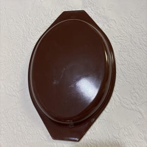 Op de afbeelding: Een bruin, ovaalvormig deksel voor een ovenschaal. Het deksel is effen, glanzend bruin van kleur en heeft een licht verhoogde rand. De schaal is ontworpen voor een ovenschaal. Het deksel is gemaakt van duurzaam materiaal.