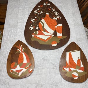 Puede incluir: Tres placas de pared de madera en forma de lágrima con incrustaciones texturizadas en naranja, blanco y marrón. Los diseños muestran bodegones que incluyen un jarrón, una piña, libros, frutas y una rama de árbol en flor.