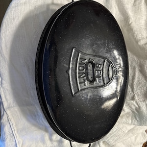 May include: Black oval-shaped cast iron casserole dish with a lid. The lid features a raised bell design with the word "MONT" and "BEL" embossed on it. The dish has two metal handles and is set on a white cloth.