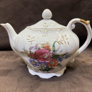 May include: A white porcelain teapot featuring a scalloped base and lid with a gold-trimmed handle and spout. The body is decorated with a colorful floral bouquet of pink, orange, blue, and yellow flowers, accented by delicate gold leaf patterns.