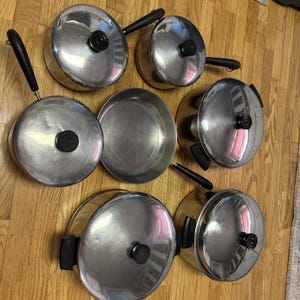 Può includere: Un set di pentole in acciaio inossidabile, tra cui pentole, padelle e coperchi. Le pentole e le padelle hanno superfici lucide e manici neri. I coperchi hanno pomelli neri. Le pentole sono disposte su una superficie di legno.