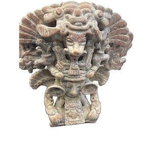 May include: A tan-coloured stone sculpture featuring a central figure with a large, elaborate headdress. The figure has prominent circular ear ornaments and a detailed chest piece. The upper portion of the sculpture displays a stylised animal face.