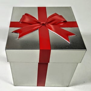 Op de afbeelding: Een vierkante zilverkleurige metalen geschenkdoos met een felrood lintontwerp dat over de bovenkant en zijkanten is gedrukt. Het deksel is versierd met een opvallende, glanzende rode strik die in reliëf is aangebracht voor een 3D-effect.