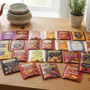 Könnte beinhalten: Eine Sammlung verschiedener Twinings-Teebeutel, die auf einer Holzoberfläche angeordnet sind. Die rechteckigen Packungen zeigen diverse farbenfrohe Designs, Markenlogos und Geschmacksbezeichnungen wie Chai, Zitrone, Wildbeere und Kakao.
