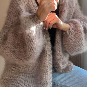 Op de afbeelding: Een lichtbruin, oversized gebreid vest met lange, volumineuze mouwen. De open trui heeft een zachte, donzige textuur en een relaxte pasvorm. Het vest heeft een neutrale kleur, geschikt om in laagjes te dragen en voor dagelijks gebruik.
