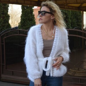 Op de afbeelding: Een witte, pluizige gebreide cardigan met een strikceintuur. De open trui heeft lange mouwen en wordt gecombineerd met een blauwe jeans. Een gouden ketting en een zwarte zonnebril maken de look compleet. De cardigan is een gezellige, stijlvolle buitenkleding.