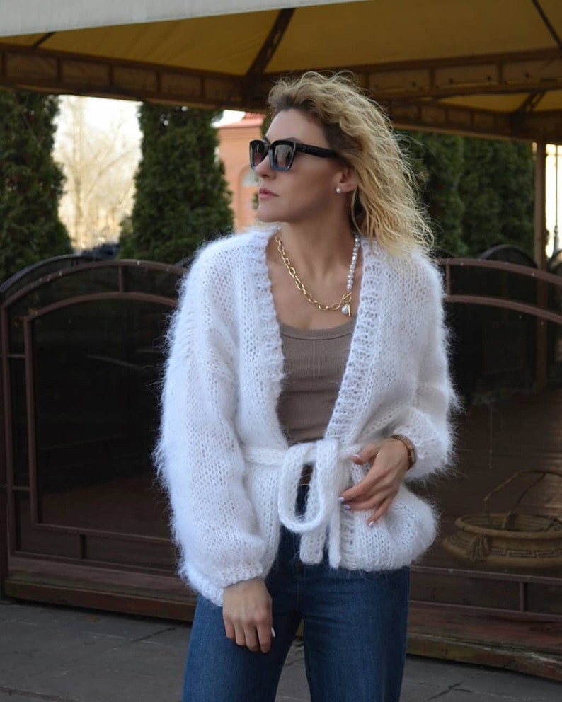 Op de afbeelding: Een witte, pluizige gebreide cardigan met een strikceintuur. De open trui heeft lange mouwen en wordt gecombineerd met een blauwe jeans. Een gouden ketting en een zwarte zonnebril maken de look compleet. De cardigan is een gezellige, stijlvolle buitenkleding.