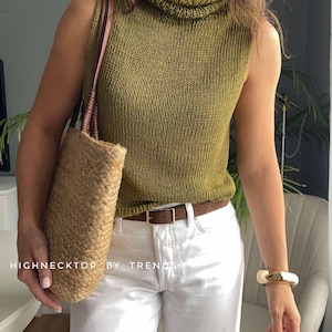 Puede incluir: Un top de punto sin mangas en color verde oliva con cuello alto, combinado con pantalones blancos, un cinturón de cuero marrón con hebilla dorada, un bolso de paja trenzada con asas de cuero marrón y un brazalete dorado. El texto dice: HIGHNECKTOP_BY_TRENDS.