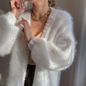 Op de afbeelding: Een lange, open witte cardigan met volumineuze mouwen. De cardigan wordt gecombineerd met een gouden ketting. Het breiwerk heeft een zachte, donzige textuur. De outfit wordt gecompleteerd met een gele top en een donkere broek.