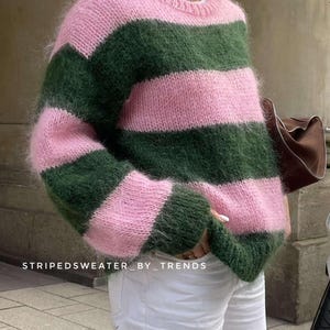 Peut inclure: Un pull à rayures roses et vertes. Le pull présente des rayures horizontales roses et vertes. Le texte "STRIPEDSWEATER_BY_TRENDS" est visible en bas de l'image. Il est porté avec un pantalon blanc.