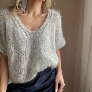 Peut inclure: Un pull en maille gris clair, moelleux, à col en V et manches courtes. Le pull est fait d'un tissu doux et léger et a une coupe décontractée.