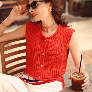 May include: A sleeveless, red knitted cardigan with gold buttons. The cardigan is worn with white trousers and sunglasses. The person is seated at a table with a glass of iced coffee. The overall theme is casual summer fashion.
