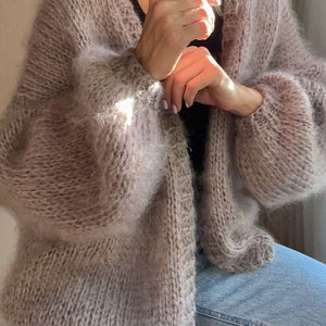 以下が含まれることがあります： ゆったりとしたシルエットと長袖の、柔らかなライトブラウンのカーディガン。ふわふわの糸でできており、少しざっくりとした編み地です。