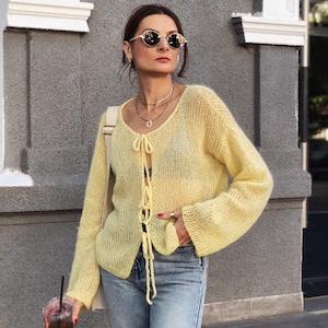 Peut inclure: Un cardigan en tricot jaune clair à devant ouvert, avec des manches longues et une fermeture à nouer. Le cardigan est porté avec un jean bleu et des lunettes de soleil. Un sac à bandoulière et une boisson dans un gobelet transparent avec une paille sont également visibles.