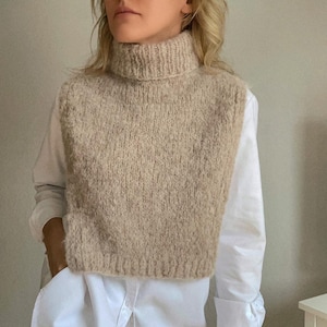 Peut inclure: Un gilet en tricot beige à col roulé avec une texture duveteuse. Le gilet est court et a un col haut.