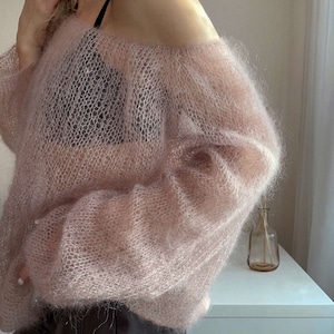 Op de afbeelding: Een lichtroze, open geweven trui met lange mouwen en een off-the-shoulder ontwerp. De afbeelding toont de textuur van de trui en hoe deze valt. De trui wordt gedragen met een zwarte top. Delicate breiwerk.