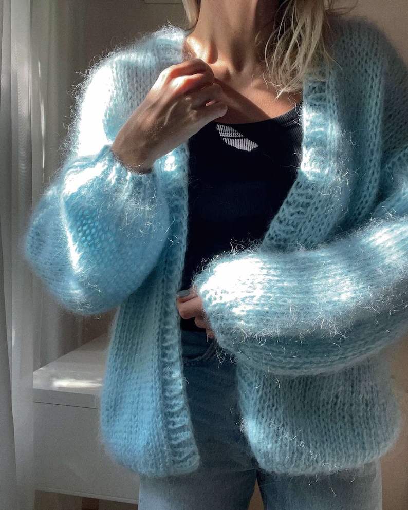 Op de afbeelding: Een lichtblauwe cardigan met open voorkant en lange, pofmouwen. De gebreide trui heeft een zachte, fuzzy textuur en wordt over een zwarte top gedragen. De cardigan is licht pastelblauw.
