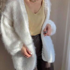 Op de afbeelding: Een witte, openvallende cardigan met een pluizige, getextureerde brei. De trui met lange mouwen wordt gecombineerd met een gele camisole en een bruine broek. Een gouden ketting voegt een vleugje elegantie toe.