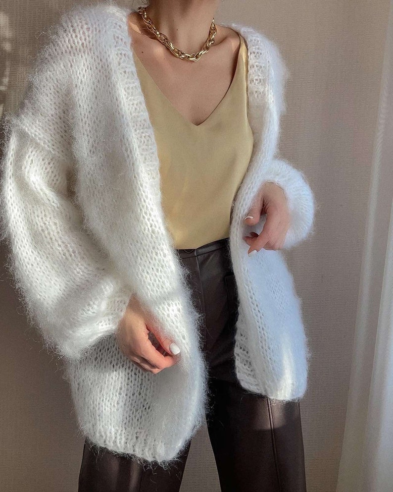 Op de afbeelding: Een witte, openvallende cardigan met een pluizige, getextureerde brei. De trui met lange mouwen wordt gecombineerd met een gele camisole en een bruine broek. Een gouden ketting voegt een vleugje elegantie toe.