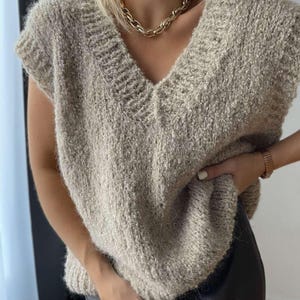 May include: A beige, sleeveless sweater with a V-neck. The knitwear has a textured appearance. A gold chain necklace is worn with the sweater. The sweater is paired with dark brown leather pants. A gold watch is worn on the wrist.