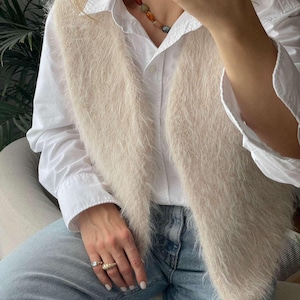 May include: A beige, fluffy vest worn over a white button-down shirt and blue jeans. The vest has a soft, textured appearance. A multi-coloured beaded necklace and gold rings are also visible. The overall style is casual and comfortable.