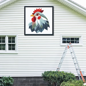 May include: A framed artwork featuring a geometric rooster design. The rooster's head is depicted with red, blue, and gray shapes against a white background. The artwork is mounted on a white exterior wall, with a ladder and greenery visible below.