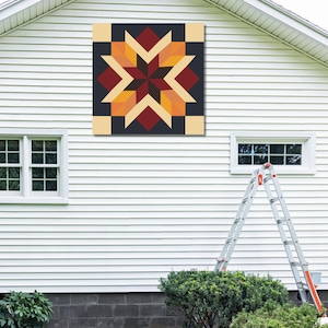 Könnte beinhalten: Ein großer, quadratischer Quiltblock mit einem Sternmuster ist an einem weißen Haus angebracht. Der Quiltblock weist ein geometrisches Muster in den Farbtönen Burgunderrot, Orange, Gelb und Schwarz auf. Das Haus hat weiße Verkleidungen, Fenster und eine Leiter.