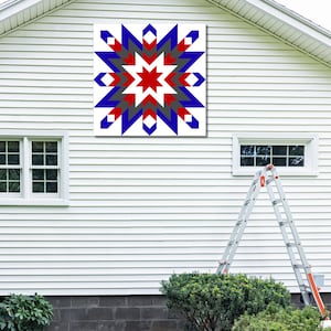 May include: A large, square, star-shaped quilt block in red, white, blue, and gray hues is mounted on the side of a white house. The star design features a central red star, surrounded by layers of blue, gray, and white geometric shapes.