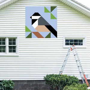 May include: A geometric bird art piece with a light blue background is mounted on a white house. The bird is depicted with black, white, grey, and tan shapes. Green and brown geometric shapes are also present. A ladder is leaning against the house.