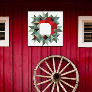 May include: A square wall art piece featuring a geometric wreath design with red, green, and brown triangles. The artwork hangs on a red wooden wall, with a vintage wooden wagon wheel below.