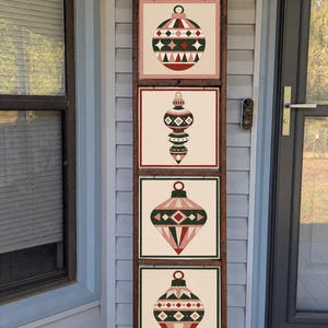 May include: A tall, dark brown wooden ladder display featuring four square panels arranged vertically. Each panel displays a unique, stylized Christmas ornament illustration in shades of pink, dark green, white, and red with geometric patterns.