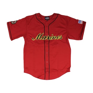 May include: Red baseball jersey with black trim and a gold "Marines" inscription. The jersey has a small American flag patch on the right sleeve and a military emblem patch on the left sleeve.