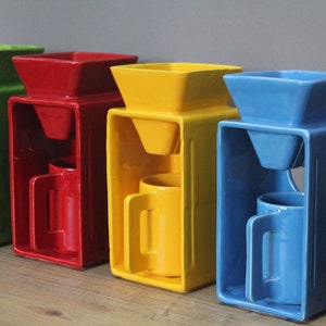 Pode incluir: Quatro cafeteiras de cerâmica em diferentes cores: verde, vermelho, amarelo e azul. Cada cafeteira tem uma base quadrada, um filtro em forma de funil e uma caneca.