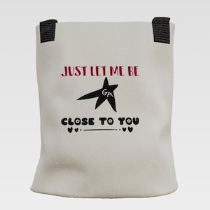 Op de afbeelding: Een lichtbeige draagtas met twee zwarte handvatten. De voorzijde is voorzien van de tekst JUST LET ME BE in het rood, een zwarte ster met de letters GA erin, en de tekst CLOSE TO YOU in het zwart met kleine bloem- en hartsymbolen eronder.
