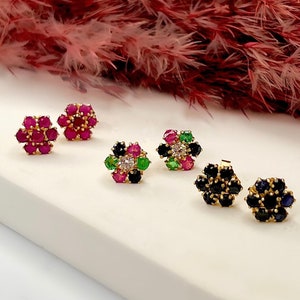 May include: Four pairs of flower-shaped earrings with different colored gemstones. The earrings are on a white surface. The earrings are made of gold and have a variety of gemstones, including red, green, and blue.
