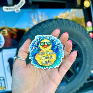 May include: A yellow rubber duck wearing sunglasses with the text "My ducks were in a row... But then I hit a curb."  The duck is surrounded by a blue and green watercolor-like design.