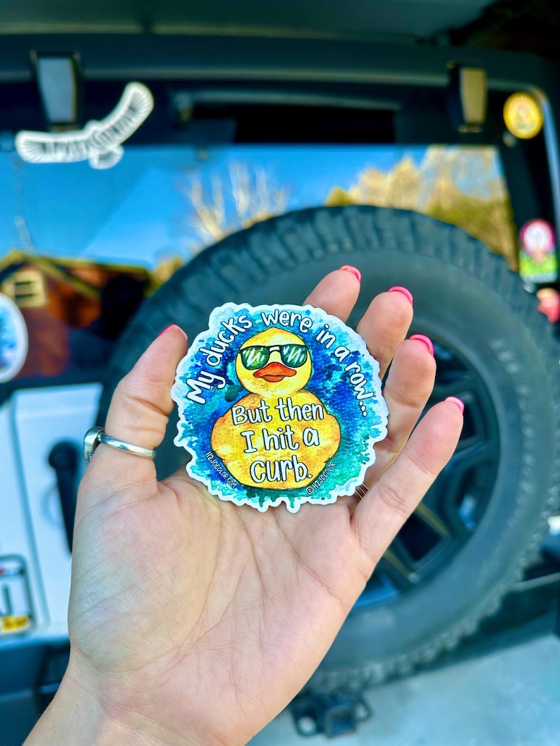 May include: A yellow rubber duck wearing sunglasses with the text "My ducks were in a row... But then I hit a curb."  The duck is surrounded by a blue and green watercolor-like design.