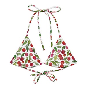 May include: A white bikini top with a watercolor cherry pattern. The top features red cherries and green leaves on a white background. The top ties at the neck and back with white straps.