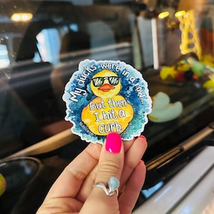 May include: A yellow rubber duck with sunglasses and a blue watercolor background. The duck has text that reads "My ducks were in a row... But then I hit a curb."