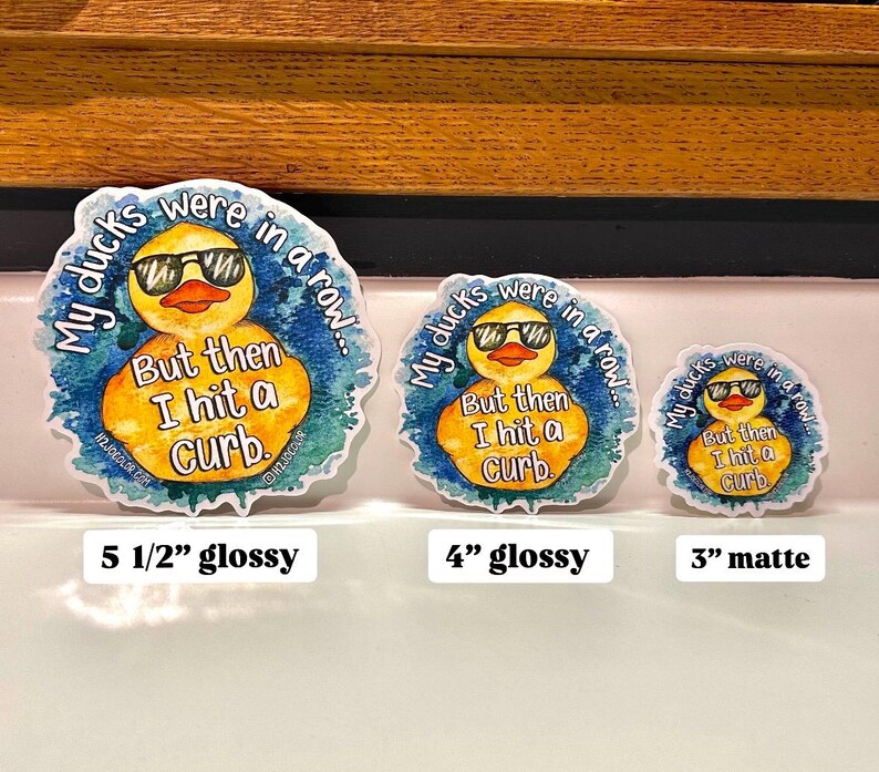May include: Three yellow rubber duck stickers with a watercolor background. The ducks are wearing sunglasses and the text reads "My ducks were in a row... But then I hit a curb." The stickers are available in three sizes: 5 1/2 inches, 4 inches, and 3 inches. The 5 1/2 inch and 4 inch stickers are glossy, while the 3 inch sticker is matte.