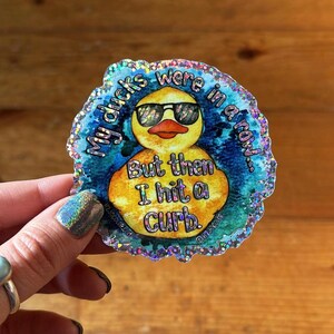 May include: A glittery sticker with a yellow rubber duck wearing sunglasses. The text on the sticker reads "My ducks were in a row... But then I hit a curb."