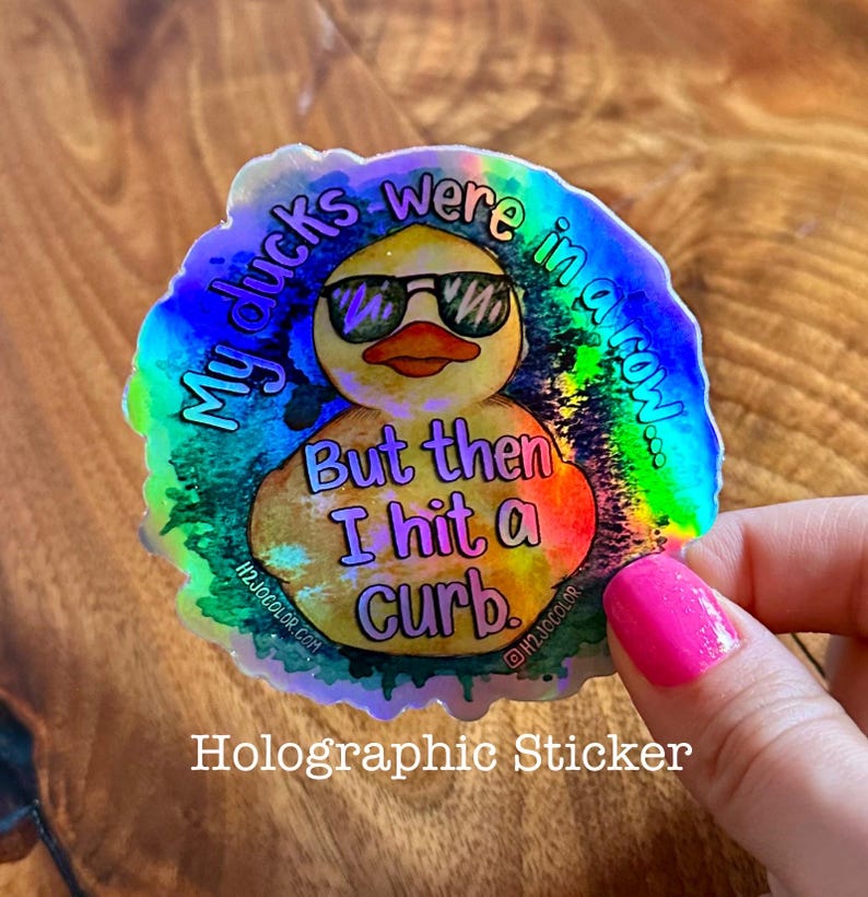 May include: A holographic sticker with a yellow rubber duck wearing sunglasses. The text on the sticker reads "My ducks were in a row... But then I hit a curb."