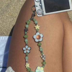 May include: A beaded camera strap featuring green beads and white flower charms. The strap is attached to a silver digital camera. The flowers have white petals and copper centers. The strap also has small yellow star-shaped charms.