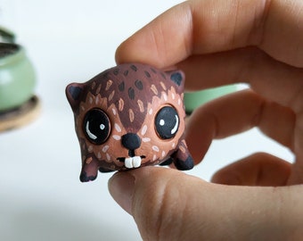 May include: A small, brown and black hand-painted clay figurine of a cartoon animal with large eyes and a toothy grin.