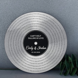 May include: A silver-colored, circular decorative piece with the words "Can't Help Falling in Love" and the names "Carly & Jordan" in a central black circle. The date "02-28-2020" is below the names. The lyrics of the song are printed around the edge.