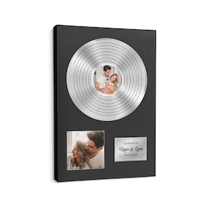 May include: A black framed canvas print featuring two photos and a silver-colored circular design with text. The top photo shows a man holding a child. The bottom photo shows a couple. The text on the print reads "Look At Us" and the names "Roger & Lynn" with the date "09.27.2014".