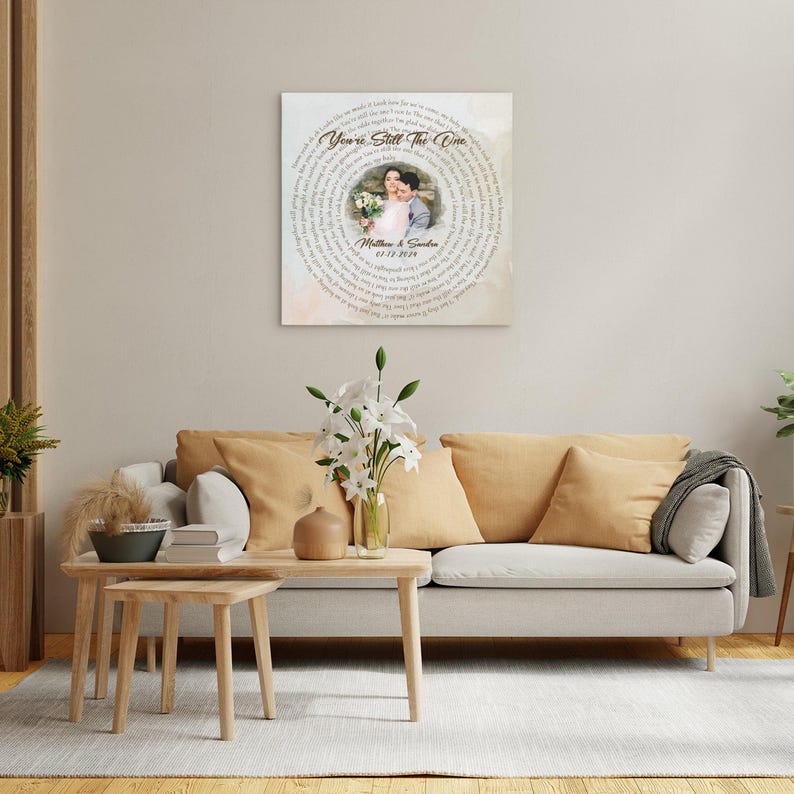 May include: A square canvas print featuring a couple's photo surrounded by song lyrics. The text reads "You're Still The One" with the couple's names and wedding date below. The print is displayed above a beige sofa.