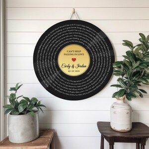 May include: A circular wall hanging designed like a vinyl record with a black outer ring featuring the lyrics to Can't Help Falling in Love. The center gold label displays the text CAN'T HELP FALLING IN LOVE, a red heart, Carly & Jordan, and 02-28-2020.