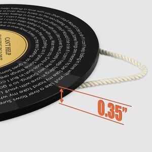 May include: A circular black wall plaque designed like a vinyl record with lyrics from Can't Help Falling in Love printed in white text. It features a gold center label and a white rope for hanging. A graphic indicates a thickness of 0.35 inches.