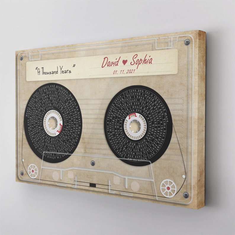 May include: A vintage-style canvas print resembling a cassette tape. The print features the song title "A Thousand Years" and the names "David" and "Sophia" with a date. The cassette reels are black with song lyrics, and the overall color scheme is beige and brown.